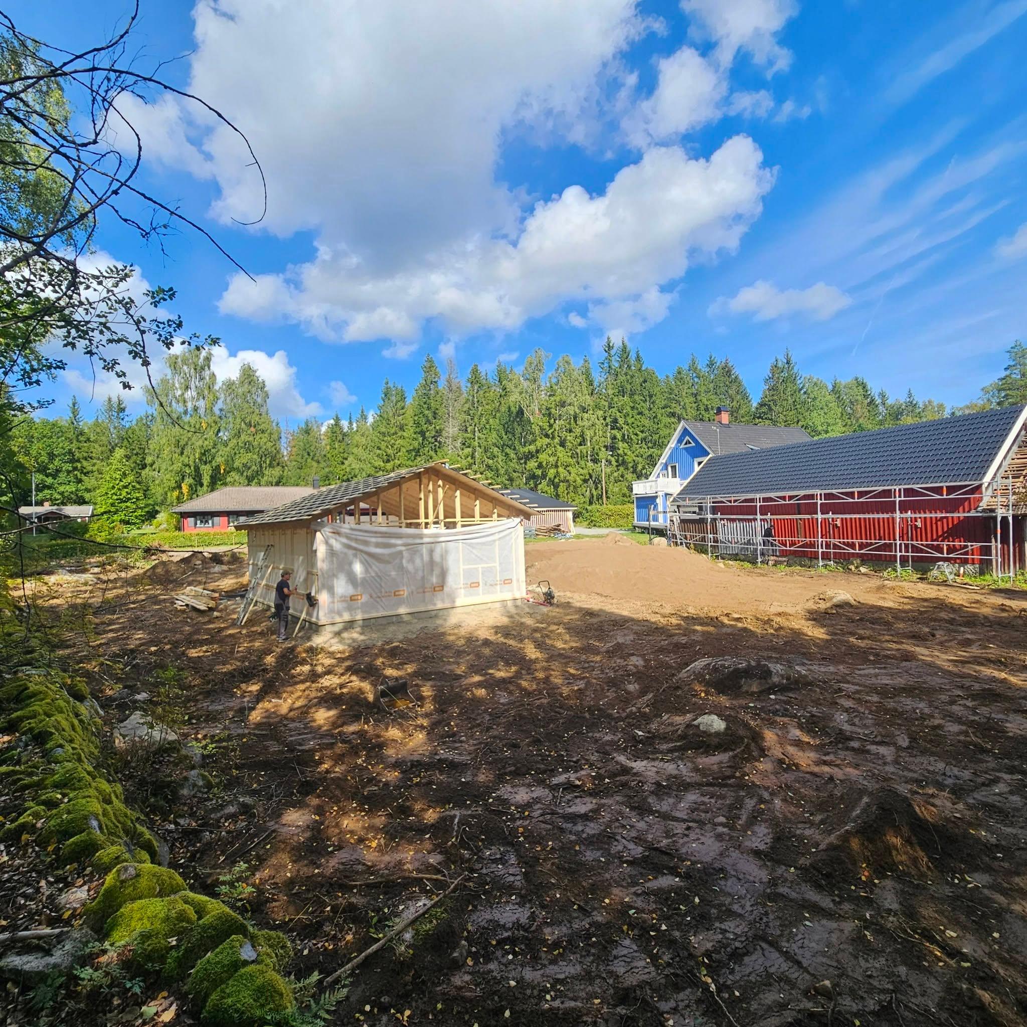 Nybyggt attefallshus i trä, planerad gårdsplan och en lada med ställning i bakgrunden